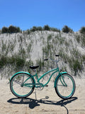 Bella mint green beach bicycle on a sandy beach with grassy dunes and clear blue sky.